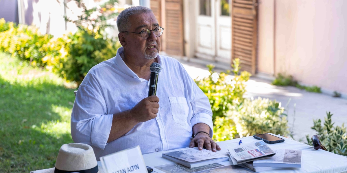 Beyaz gömlekli, gözlüklü ve kısa saçlı orta yaşlı bir erkek, dış mekanda bir masa başında elinde mikrofonla konuşma yapıyor. Masanın üzerinde birkaç kitap, bir gazete, bir akıllı telefon ve bir şapka bulunuyor. Arka planda yeşil bitkiler ve aydınlık bir bina girişi yer alıyor.