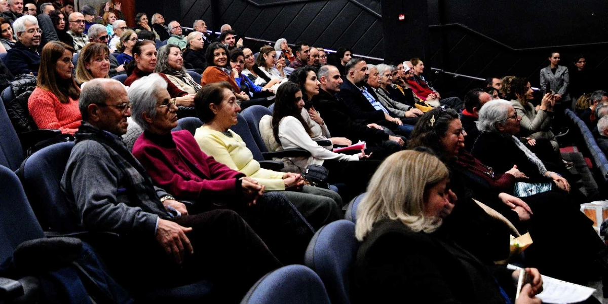 Kapalı bir konferans salonunda, mavi koltuklarda yan yana oturmuş, dikkatle sahneye doğru bakan kalabalık bir izleyici grubu. Farklı yaş gruplarından oluşan katılımcılar, Uğur Mumcu'yu anma etkinliği kapsamında bir araya gelmiş, ciddiyetle etkinliği takip ediyor.