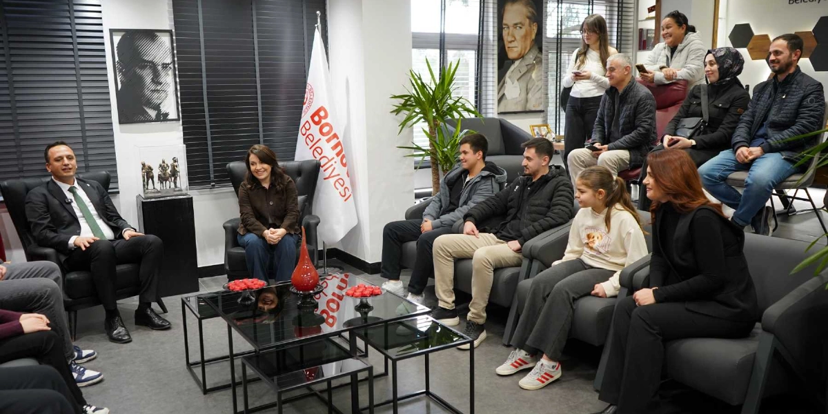 Bornova Belediye Başkanı, makam odasında bir grup genç ve yetişkinle bir araya gelerek sohbet ediyor. Odanın duvarında Mustafa Kemal Atatürk'ün portresi ve Bornova Belediyesi bayrağı yer alıyor. Ortada siyah camlı bir sehpa üzerinde ikramlıklar bulunurken, katılımcılar karşılıklı koltuklarda oturuyor.