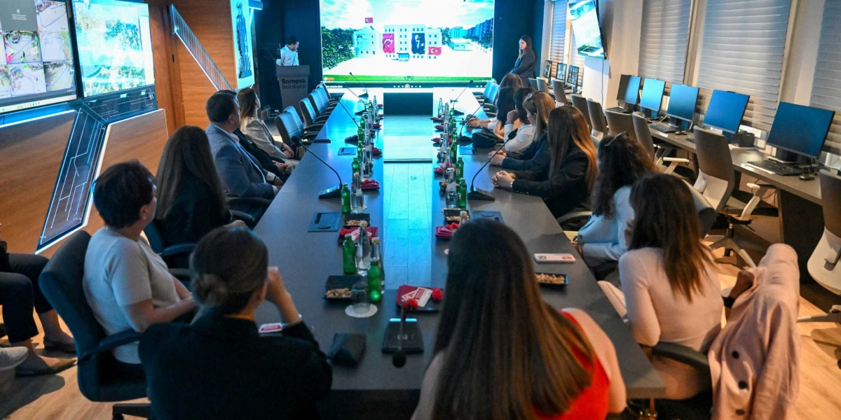 Bornova Belediyesi'ne ait modern bir toplantı salonunda, uzun bir masa etrafında toplanmış bir grup kadın ve erkek katılımcı yer alıyor. Salonun ön tarafındaki dev ekranda, üzerinde Türk bayrağı ve Atatürk posteri bulunan bir bina görseli yansıtılıyor. Masanın üzerinde mikrofonlar, su şişeleri ve atıştırmalıklar bulunurken, yan duvarlarda güvenlik kameralarını gösteren çoklu ekranlar ve çalışma istasyonları yer alıyor. Katılımcılar, ekranı ve toplantıyı dikkatle takip ediyor.