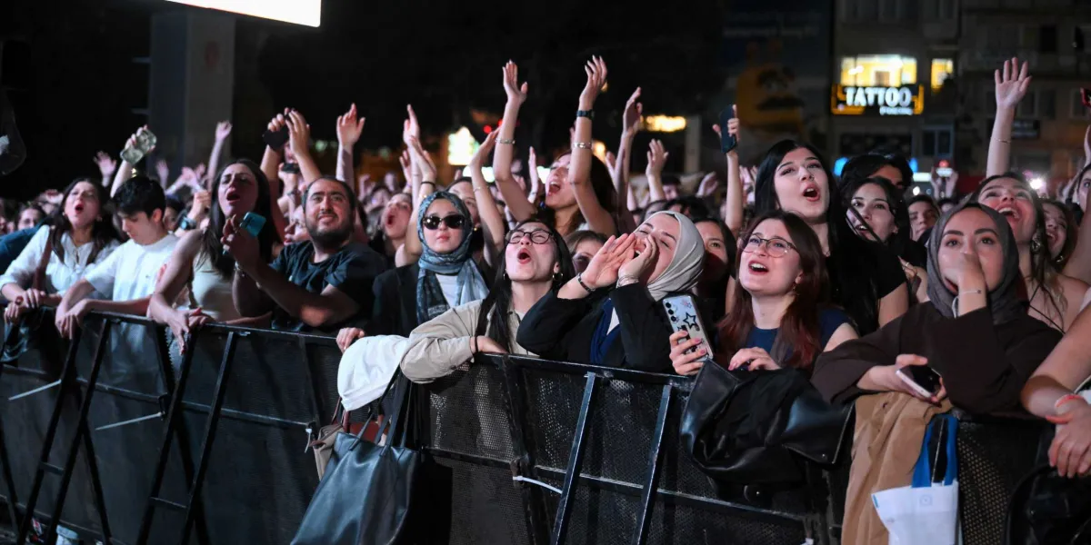Gece vakti düzenlenen bir açık hava konserinde, metal bariyerlerin önünde toplanmış, ellerini havaya kaldırmış ve coşkuyla şarkı söyleyen kalabalık bir genç grubu. Ön planda, bazıları başörtülü olan genç kadınlar ve erkekler heyecanla sahneye doğru bakıyor, kimileri ellerindeki telefonlarla çekim yapıyor. Arka planda ise şehir ışıkları ve üzerinde 