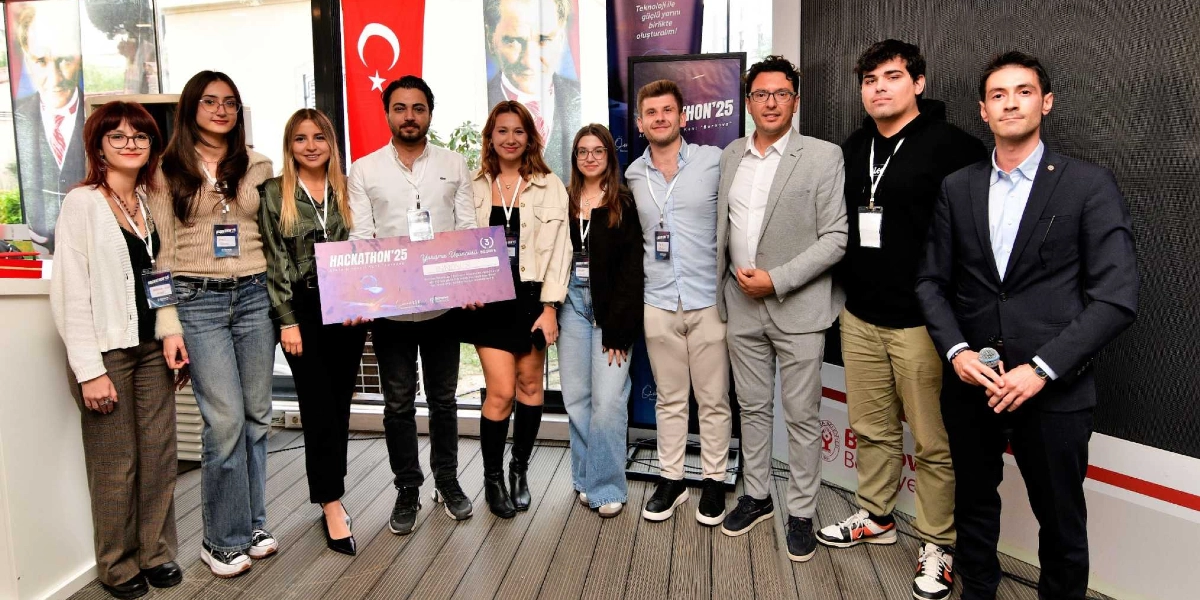 Hackathon'25 etkinliğinde ödül kazanan bir grup genç ve organizasyon yetkilileri yan yana duruyor. Grubun ortasındaki iki kişi, üzerinde 