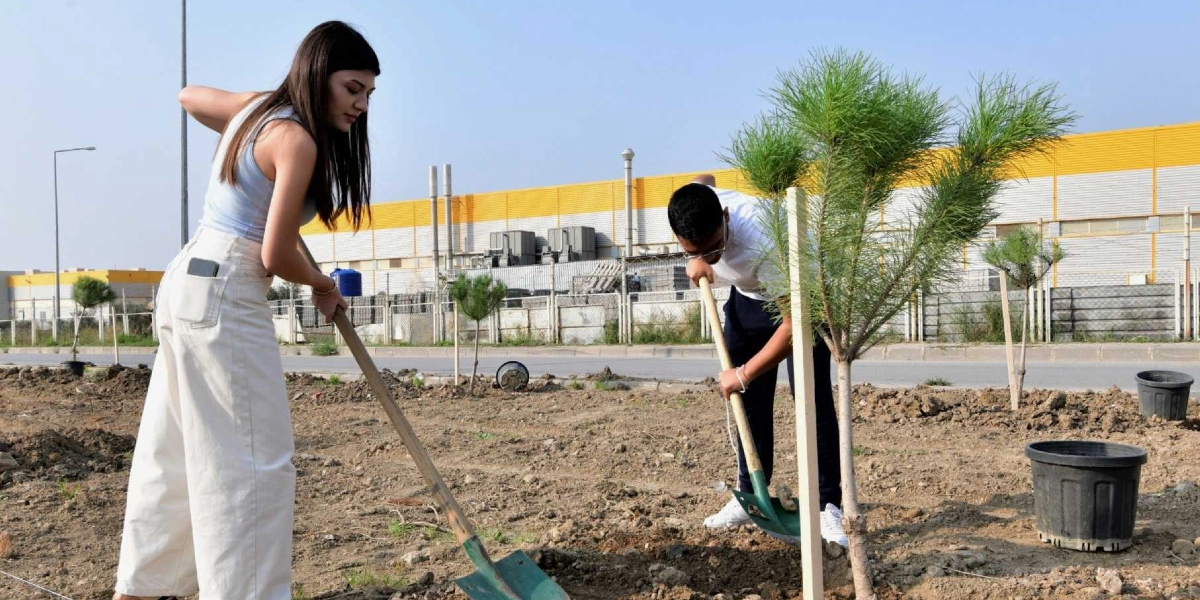 Açık havada, toprak bir alanda fidan dikimi yapan iki genç; sol tarafta açık renkli kıyafetler giymiş genç bir kadın, sağ tarafta ise beyaz tişörtlü bir genç erkek ellerinde küreklerle toprağı kazıyor. Arka planda sarı ve gri renkli büyük bir endüstriyel yapı ile yol kenarındaki fidanlar görülüyor.