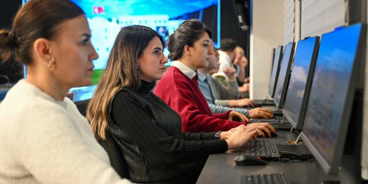 Modern bir ofis ortamında bilgisayar başında çalışan üç kadın ve bir erkek, önlerindeki ekranlara odaklanmış şekilde klavye ve fare kullanarak dijital veriler üzerinde çalışıyor. Arka planda ise büyük bir ekranda Türkiye bayrağı ve çeşitli grafiklerin yer aldığı bir sunum yansıtılıyor.