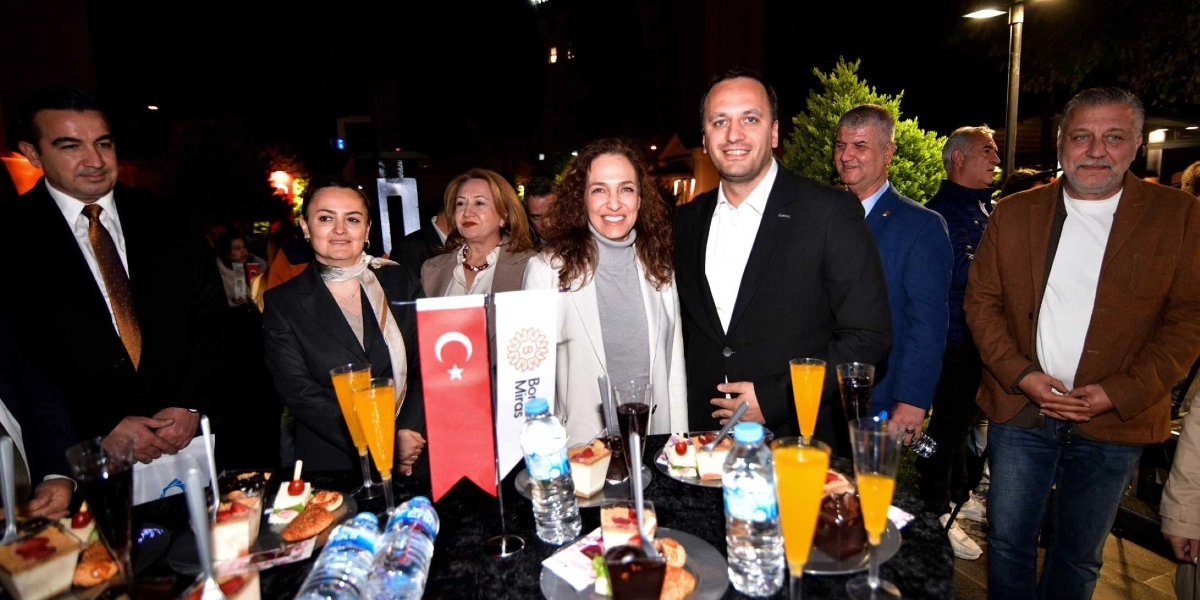 Gece vakti dış mekanda düzenlenen bir etkinlikte, siyah örtülü bir masanın etrafında duran bir grup insan gülümsüyor. Masanın üzerinde Türk bayrağı, Bornova Belediyesi'ne ait 