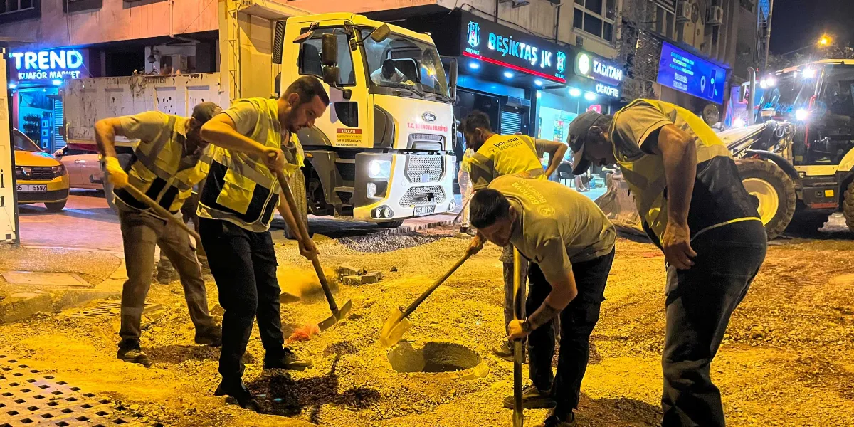 Gece vakti Bornova Belediyesi üniformalı ve reflektörlü yelekli işçiler, bir cadde üzerinde kazı ve yol yenileme çalışması yapıyor. İşçilerden bazıları ellerindeki küreklerle toprağı kazarken, arkalarında T.C. Bornova Belediyesi yazılı bir kamyon ve bir iş makinesi park halinde bekliyor. Arka planda ise ışıklı tabelalarıyla 