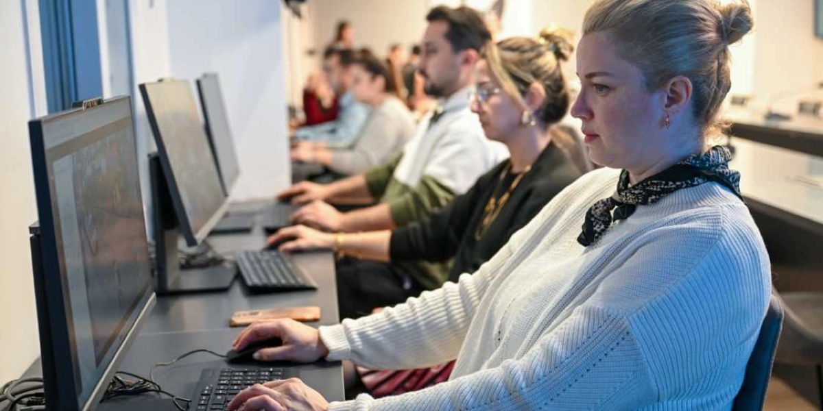 Ofis ortamında yan yana dizilmiş bilgisayarların başında çalışan bir grup insan. Ön planda, beyaz kazaklı ve boynunda desenli bir fular olan sarışın bir kadın, bilgisayar ekranına odaklanmış şekilde fareyi kullanıyor. Onun hemen arkasında, aynı hizada oturan diğer çalışanlar da dikkatle bilgisayar ekranlarını inceliyor.
