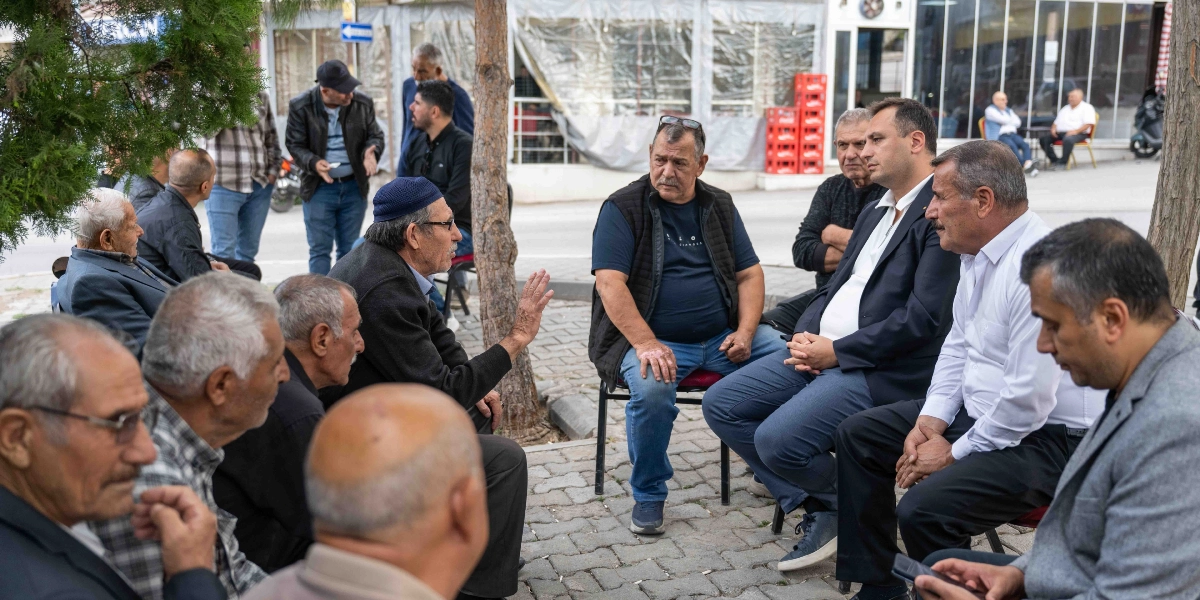 Sokakta, parke taşlı bir alanda bir araya gelmiş bir grup erkek, sandalyelerde oturarak sohbet ediyor. Grubun merkezinde, lacivert ceketli ve beyaz gömlekli bir erkek, diğerlerinin anlattıklarını dikkatle dinliyor. Yanında beyaz gömlekli bir başka erkek otururken, karşısında ise el hareketleriyle konuşan yaşlı bir adam yer alıyor. Çevredeki diğer kişiler de dikkatle konuşmaya eşlik ediyor; ortam samimi ve halkla iç içe bir toplantı havası taşıyor.