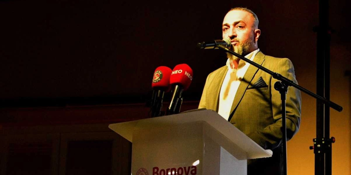 Koyu renkli bir arka plan önünde, üzerinde Bornova Belediyesi logosu bulunan bir kürsüde konuşma yapan, kirli sakallı, bej renkli ceket giymiş bir erkek. Kürsüde iki adet mikrofon bulunuyor; mikrofonlardan birinin üzerinde kırmızı renkli 