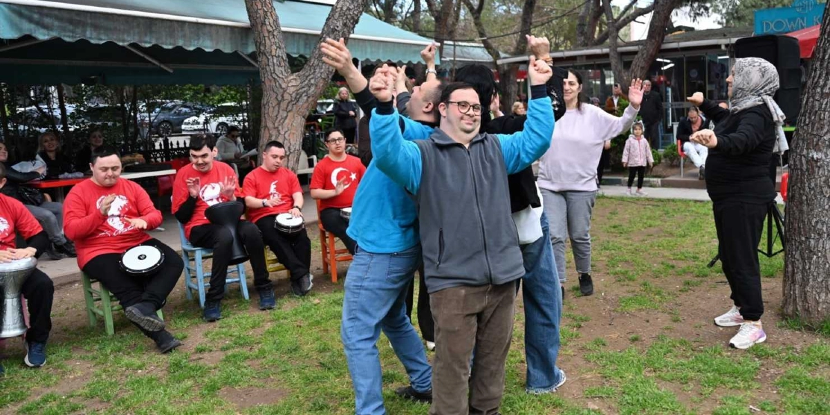 Açık havada, ağaçların altında bir araya gelmiş bir grup insan müzik eşliğinde eğleniyor. Ön planda, mavi sweatshirt ve kahverengi pantolon giyen, gözlüklü bir genç, kollarını havaya kaldırarak neşeyle dans ediyor. Arkasında kırmızı tişörtlü bir grup genç, ellerinde darbuka ve tef gibi vurmalı çalgılarla ritim tutuyor. Sağ tarafta başörtülü bir kadın ve açık renkli kıyafetli bir başka kadın da dansa eşlik ediyor. Arka planda kafe binası, masalar ve etkinliği izleyen diğer insanlar görülüyor.