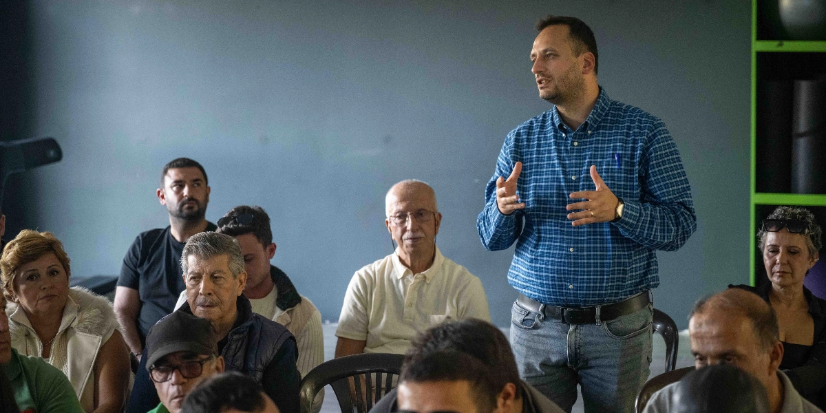 Mavi ekose gömlekli bir erkek, kapalı bir mekânda toplanmış bir gruba hitaben konuşma yapıyor. Konuşmacı ellerini kullanarak açıklamalarda bulunurken, önünde oturan farklı yaş gruplarından oluşan katılımcılar dikkatle onu dinliyor.