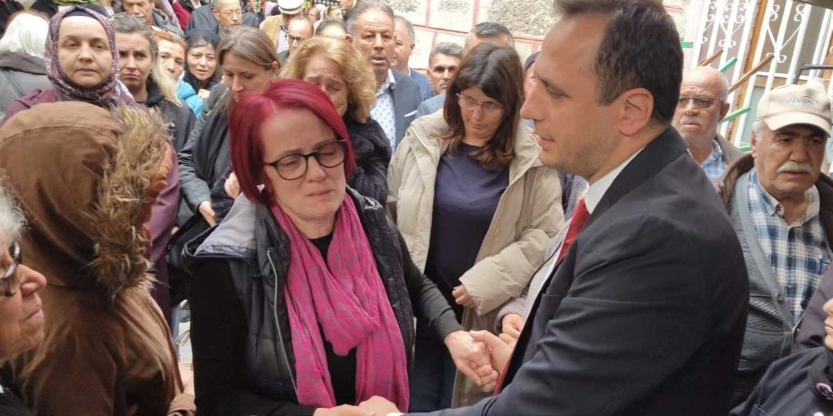 Kalabalık bir ortamda, siyah kıyafetli ve pembe şallı, gözlüklü bir kadın üzgün bir ifadeyle elini uzatmış, koyu renk takım elbiseli ve kırmızı kravatlı bir erkekle tokalaşıyor. Çevredeki insanlar da yaslı ve hüzünlü bir ifadeyle onları izliyor.