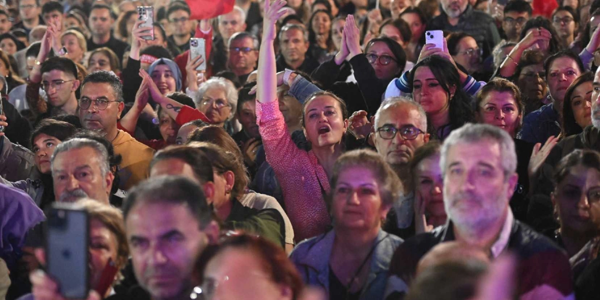 Bornova'da düzenlenen 29 Ekim Cumhuriyet Bayramı kutlamalarında, ellerinde telefonlarıyla çekim yapan ve coşkuyla eşlik eden kalabalık bir insan grubu. Ön planda bir kadın elini havaya kaldırmış şarkı söylerken, çevresindeki insanlar dikkatle sahneye bakıyor.