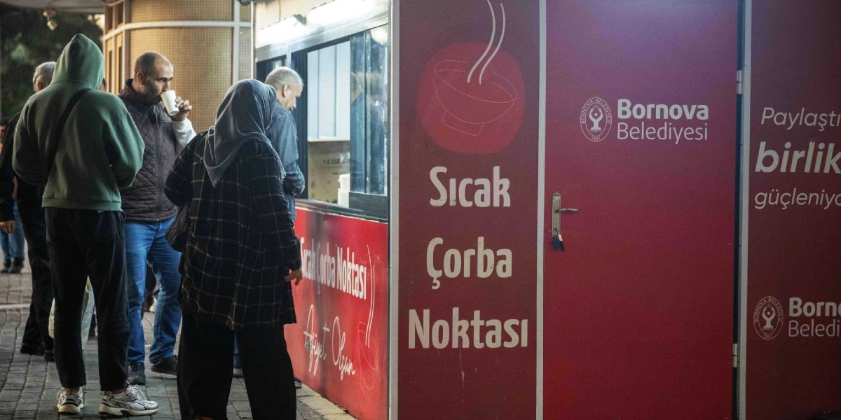 Bornova Belediyesi'ne ait kırmızı renkli bir çorba dağıtım noktasının önünde, ellerinde karton bardaklarla çorbalarını içen ve sıra bekleyen insanlar görülüyor. Kabinin üzerinde 