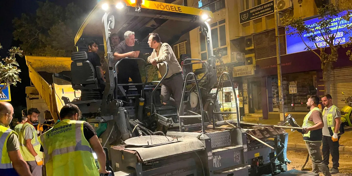 Gece vakti Bornova Belediyesi ekiplerinin yürüttüğü yol asfaltlama çalışması. Sarı renkli bir Vögele asfalt serici makinesinin üzerinde üç kişi dururken, çevresinde ise üzerinde reflektörlü yelekler bulunan belediye işçileri çalışmaktadır. Arka planda dükkanların bulunduğu bir cadde ve tabela detayları yer almaktadır.