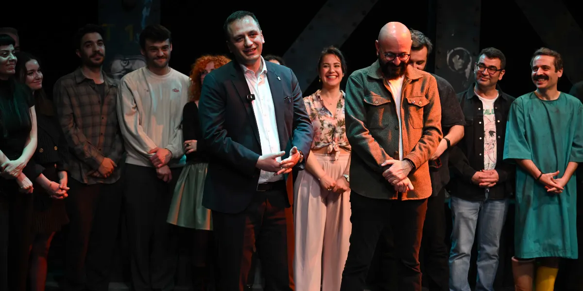 Sahne üzerinde yan yana duran, aralarında Bornova Belediye Başkanı Mustafa İduğ'un da bulunduğu bir grup tiyatro kursiyeri ve eğitmeni, seyircilere doğru gülümseyerek selam veriyor. Kursiyerler günlük kıyafetler ve bazıları kostüm giymiş şekilde sahnede yer alıyor.
