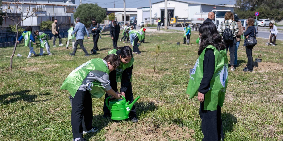 Üzerlerinde yeşil yelekler bulunan öğrenciler, açık bir arazide fidan dikimi ve sulama etkinliği gerçekleştiriyor. Ön planda iki öğrenci yeşil bir sulama kabıyla yeni dikilmiş bir fidanı sularken, arkalarında diğer öğrenciler ve yetişkinler farklı noktalarda ağaç dikmeye devam ediyor. Arka planda ise sanayi yapıları ve park halindeki araçlar görülüyor.