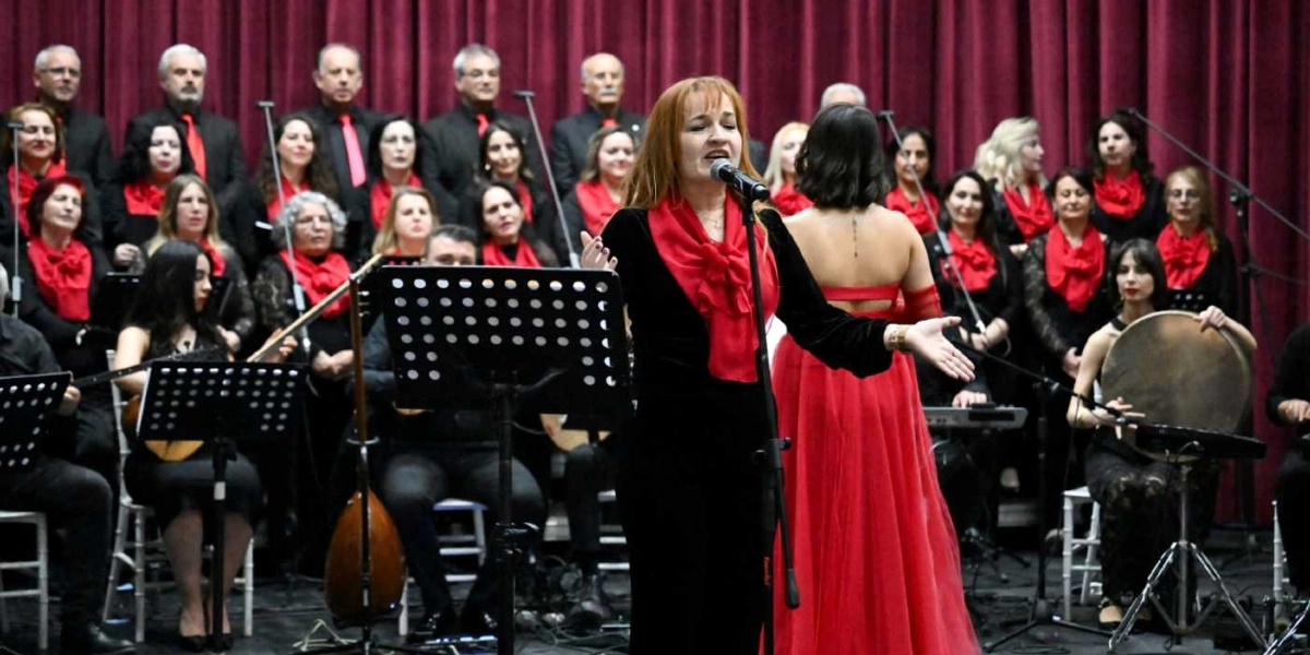 Bordo perdeler önünde sahne alan bir koro ve solist grubu. Ön planda, siyah kıyafetli ve kırmızı fularlı kadın solist mikrofon başında şarkı söylüyor. Yanında kırmızı uzun elbiseli bir başka kadın sanatçı ve sağ tarafta elinde tef tutan bir müzisyen yer alıyor. Arka planda ise siyah kıyafetler giymiş, boyunlarında kırmızı fularlar bulunan kalabalık bir koro grubu, nota sehpaları ve çeşitli müzik aletleriyle performans sergiliyor.