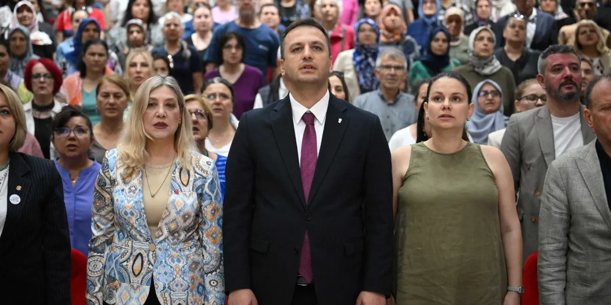 Kapalı bir salonda, ön sırada takım elbiseli ve mor kravatlı bir erkek ile yanında desenli ceketli bir kadın ve haki renkli kolsuz bir bluz giyen diğer bir kadının bulunduğu, arkalarında ise ağırlıklı olarak kadınlardan oluşan kalabalık bir grubun ciddiyetle karşıya baktığı bir toplantı anı.