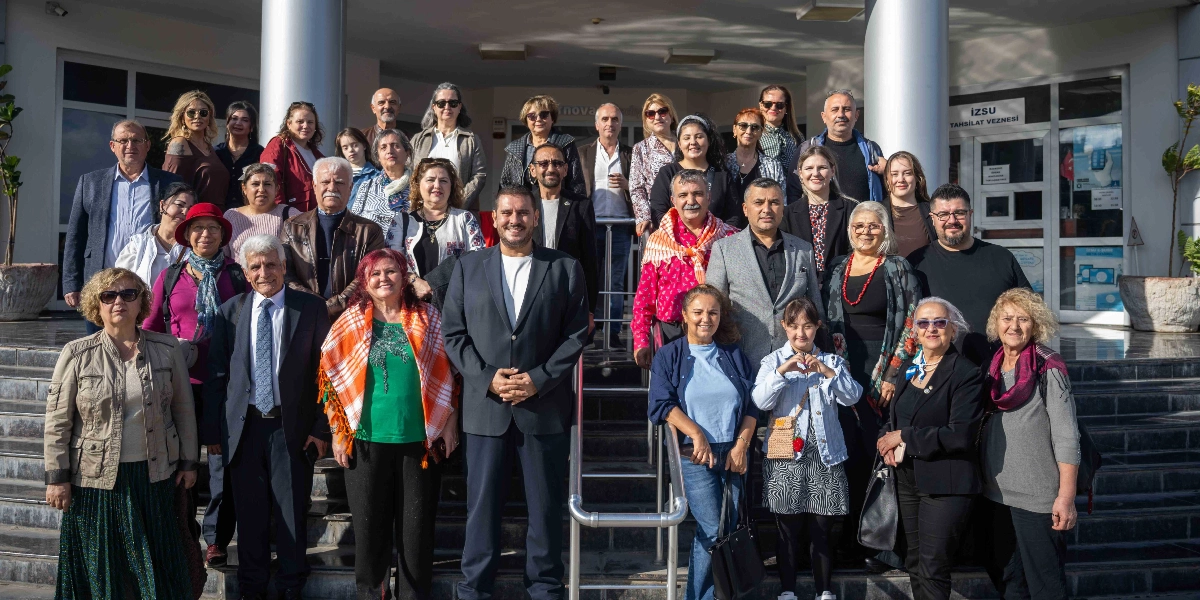 Bornova Belediyesi binasının önündeki merdivenlerde poz veren, farklı yaş gruplarından oluşan kalabalık bir grup insan. Arka planda İZSU tahsilat veznesi tabelası ve cam kapılar görülüyor. Ön planda koyu renk takım elbiseli bir erkek, yanında turuncu şallı bir kadın ve diğer katılımcılar yer alıyor. Katılımcıların çoğu gülümseyerek kameraya bakıyor, bazıları ise elleriyle kalp işareti yapıyor.