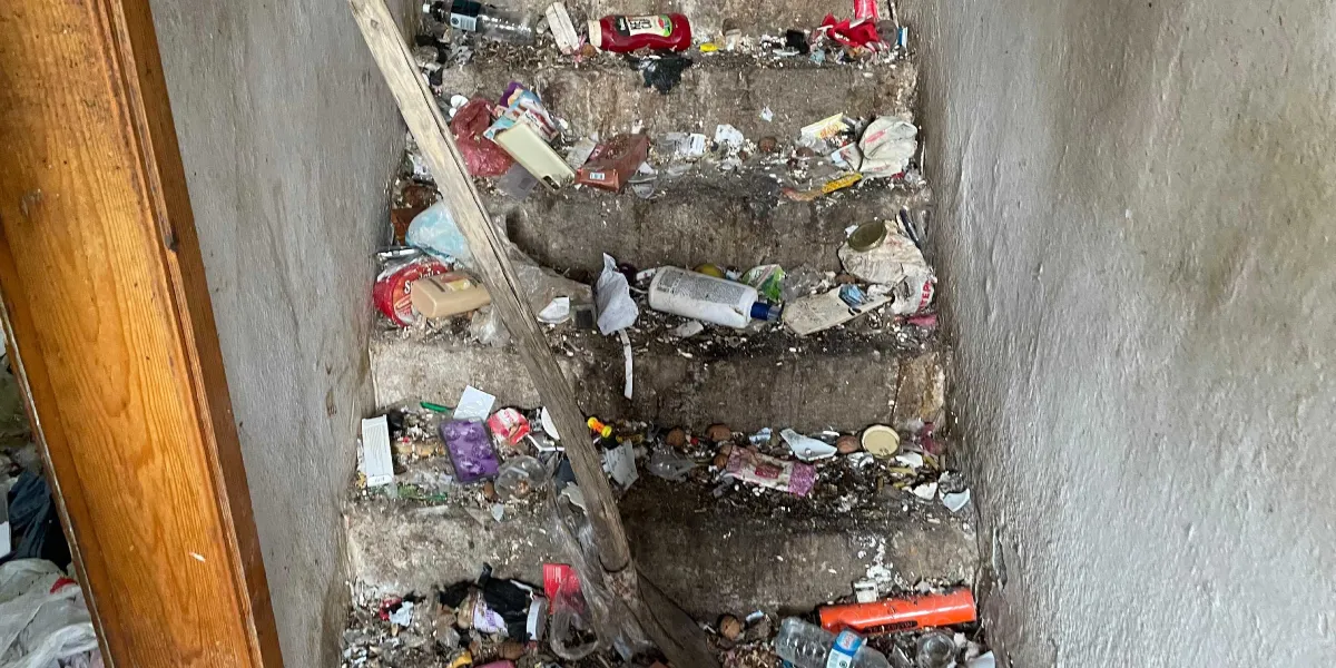 Çöp ve atıklarla dolu, bakımsız bir apartman merdiveni. Basamakların üzeri plastik şişeler, yiyecek ambalajları, kutular ve çeşitli evsel atıklarla kaplanmış durumda. Merdiven boyunca uzanan ahşap bir süpürge sapı veya destek çubuğu göze çarpıyor. Duvarlar kirli ve yıpranmış görünüyor.