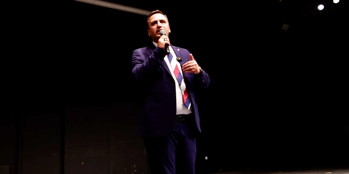 Koyu renkli bir sahnede, lacivert takım elbise ve beyaz gömlek üzerine kırmızı, beyaz ve lacivert çizgili bir kravat takmış bir erkek, elinde mikrofonla konuşma yapıyor.