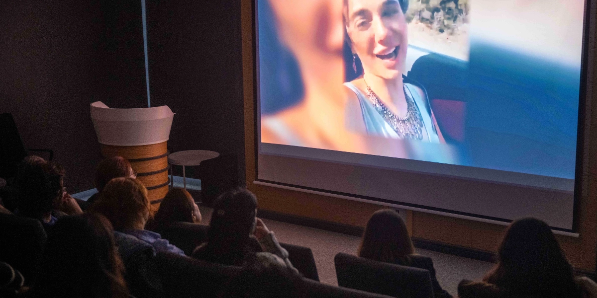 Karanlık bir konferans salonunda, beyaz perdeye yansıtılan bir videoyu izleyen bir grup insan. Perdede, dış mekanda çekilmiş, konuşan ve gülümseyen bir kadının yakın plan görüntüsü yer alıyor. Salonun sol tarafında ahşap detaylı bir kürsü bulunuyor.