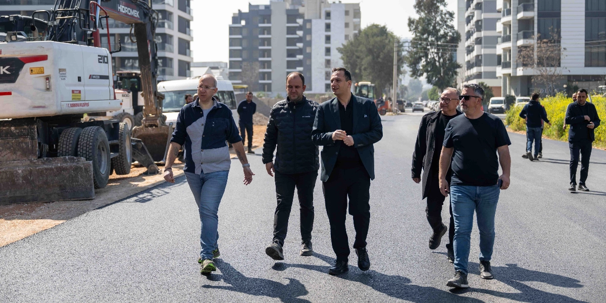 Bornova Belediye Başkanı ve beraberindeki heyet, yeni asfaltlanmış bir yolda yürüyerek çalışmaları yerinde inceliyor. Sol tarafta T.C. Bornova Belediyesi yazılı bir iş makinesi bulunurken, arka planda modern apartmanlar ve diğer görevliler yer alıyor.