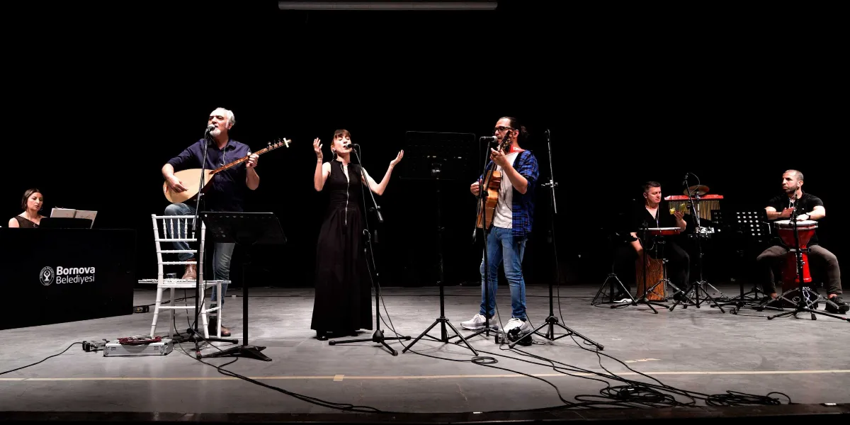 Karanlık bir sahnede, Bornova Belediyesi logolu bir kürsünün arkasında piyano çalan bir kadın ve ön planda müzik icra eden dört kişilik bir grup yer alıyor. Grupta bağlama çalan ve şarkı söyleyen bir erkek, siyah uzun elbisesiyle şarkı söyleyen bir kadın, gitar çalan bir erkek ve vurmalı çalgılarla eşlik eden iki erkek müzisyen bulunuyor. Müzisyenler mikrofonlar ve enstrümanlarıyla performans sergiliyor.