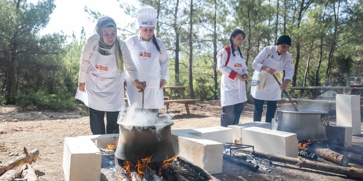 Açık havada, ağaçlık bir alanda, üzerinde Reçel Şenliği logolu beyaz önlükler giymiş dört kadın, tuğlalarla çevrili odun ateşleri üzerinde büyük metal kazanlarda reçel kaynatıyor. Kadınlardan biri başörtülü, diğeri şef şapkası takıyor ve ellerindeki kepçelerle kazanları karıştırıyorlar. Kazanlardan yoğun buhar yükseliyor.