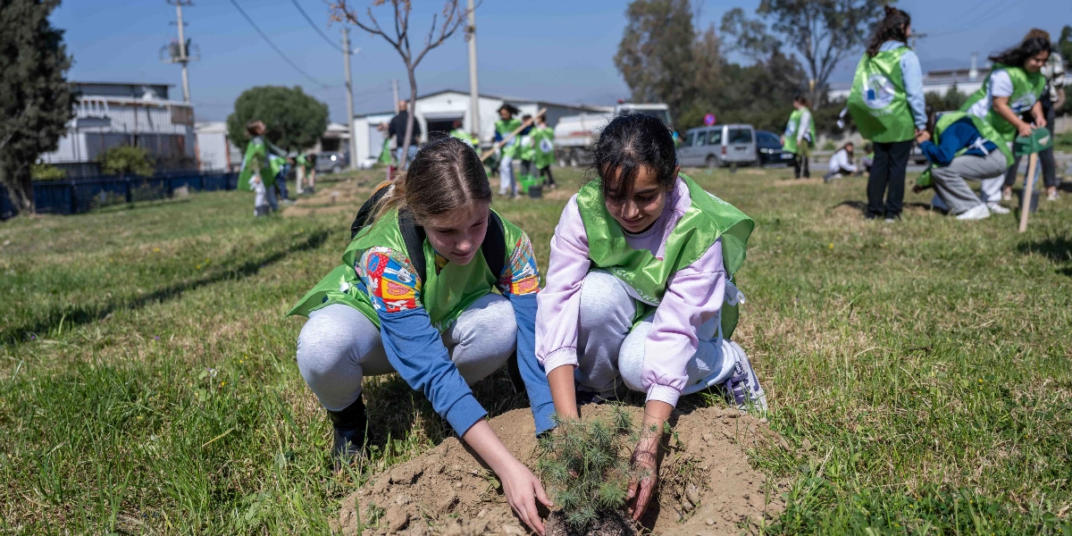 Üzerlerinde yeşil yelekler bulunan iki genç kız, açık havada bir fidanı toprakla buluşturuyor. Arka planda ise aynı yelekleri giymiş diğer çocuklar ve gençler ağaç dikme etkinliğine devam ediyor.