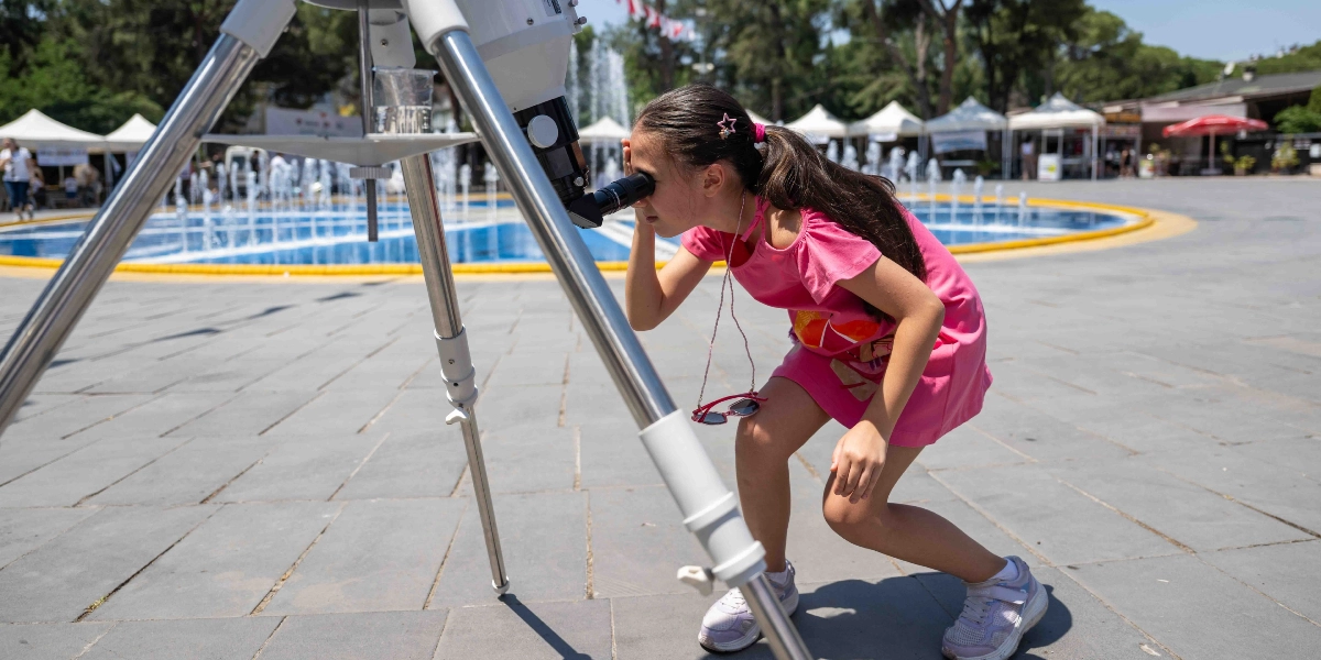 Pembe elbiseli genç bir kız, açık havada bir meydanda kurulu olan teleskobun merceğine eğilerek dikkatle bakıyor. Arka planda fıskiyeli bir havuz ve etkinlik çadırları yer alıyor.