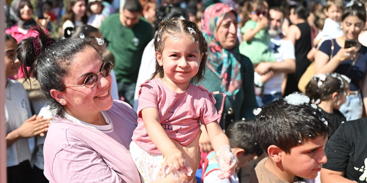 Açık havada düzenlenen bir etkinlikte, kucağındaki küçük kız çocuğuyla gülümseyerek poz veren bir kadın. Hem kadının hem de çocuğun saçlarında ve ellerinde köpükler bulunuyor. Çevrede çocukların ve ailelerin yer aldığı kalabalık bir grup, neşeli bir atmosferde etkinliğe katılıyor.