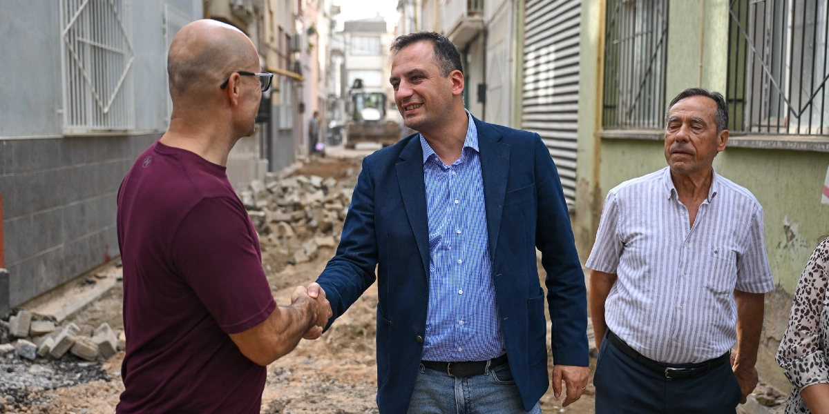 Bornova'da devam eden yol yenileme çalışmasının yapıldığı bir sokakta, lacivert ceketli ve mavi gömlekli bir yetkili, bordo tişörtlü bir vatandaşla tokalaşıyor. Yanlarında çizgili gömlekli başka bir kişi daha bulunuyor. Arka planda ise yol çalışması yapan bir iş makinesi görülüyor.