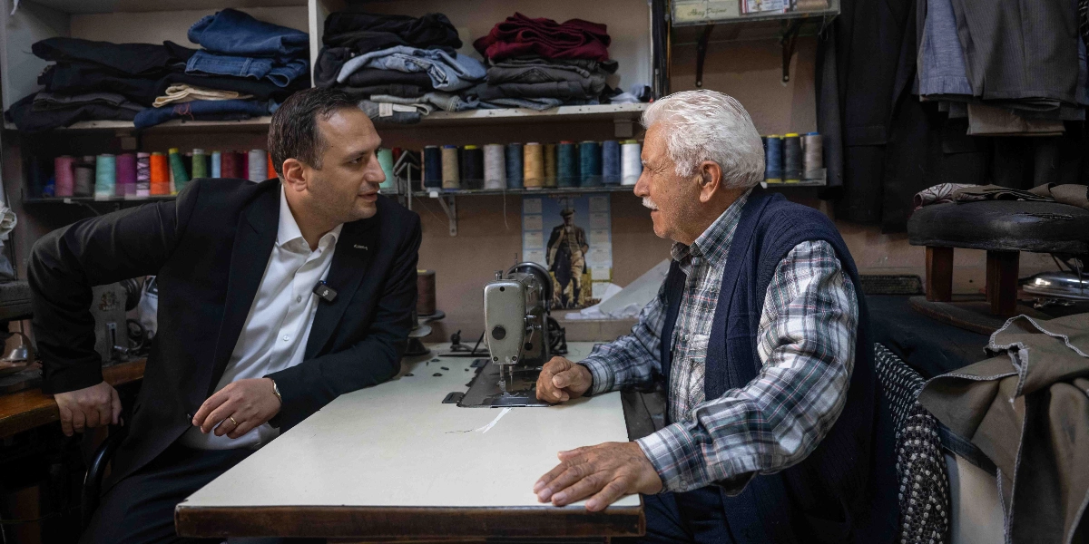 Siyah takım elbiseli bir erkek, dikiş makinesinin başında oturan beyaz saçlı, ekose gömlekli ve yelekli bir terziyle küçük bir atölyede sohbet ediyor. Arka planda raflarda dizili kumaşlar ve iplik makaraları görülüyor.