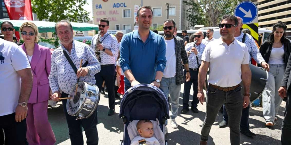 Bornova'daki bir festival yürüyüşünde, ön planda bebek arabasını süren mavi tişörtlü bir adam ve yanında beyaz tişörtlü, güneş gözlüklü bir erkek yer alıyor. Yanlarında davul çalan bir müzisyen ve arkalarında saksafon ve klarnet çalan bando ekibi bulunuyor. Grubun etrafında pembe ceketli bir kadın ve diğer katılımcılar yürürken, arka planda 