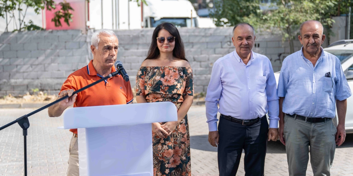 Açık havada, beyaz bir kürsü arkasında duran turuncu polo yaka tişörtlü bir adam mikrofonla konuşma yapıyor. Yanında ona eşlik eden, omuzları açık çiçek desenli bir elbise giymiş bir kadın ile açık mavi gömlekli iki erkek yan yana duruyor. Arka planda gri bir duvar ve park halindeki araçlar görülüyor.