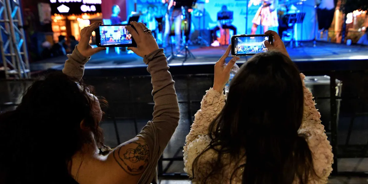 Açık havada düzenlenen bir konserde, sahneyi izleyen iki kadın ellerindeki akıllı telefonlarla çekim yapıyor. Sol taraftaki kadının omzunda dövme bulunurken, sağ taraftaki kadın açık renkli, dokulu bir üst giyiyor. Arka planda mavi ışıklarla aydınlatılmış sahnede müzisyenler yer alıyor.