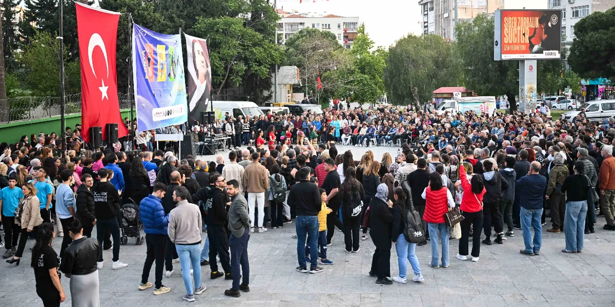 Bornova'da açık bir meydanda toplanmış kalabalık bir izleyici grubu, Dünya Dans Günü kapsamında düzenlenen gösteriyi izliyor. Meydanın arka tarafında asılı büyük bir Türk bayrağı, üzerinde dans figürleri bulunan renkli bir etkinlik afişi ve Mustafa Kemal Atatürk'ün portresinin yer aldığı bir pankart bulunuyor. Arka planda ise 
