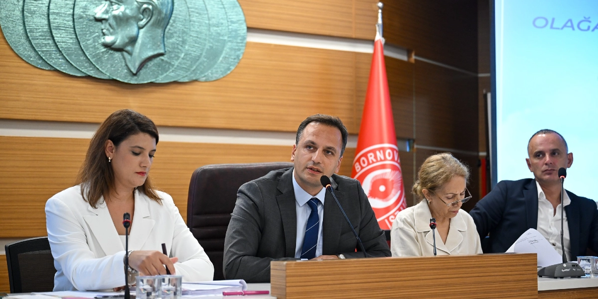 Resmi bir toplantı masasında yan yana oturan dört kişi, önlerinde mikrofonlarla konuşma yapıyor. Ortada koyu renk takım elbiseli ve mavi kravatlı bir erkek konuşurken, solunda beyaz ceketli bir kadın, sağında ise açık renkli kıyafetli bir kadın ve koyu renk ceketli bir erkek yer alıyor. Arka planda ahşap paneller üzerinde Atatürk kabartması ve Bornova Belediyesi'nin kırmızı beyaz bayrağı bulunuyor.