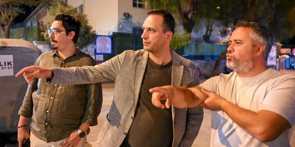 Gece vakti dış mekanda yan yana duran üç erkek, bir noktayı işaret ederek bir şeyler konuşuyor. Soldaki gözlüklü ve haki renkli gömlekli kişi, ortadaki gri ceketli kişi ve sağdaki beyaz tişörtlü kişi aynı yöne doğru bakıp elleriyle bir yeri gösteriyor. Arka planda aydınlatılmış bir bina ve çöp konteyneri bulunuyor.
