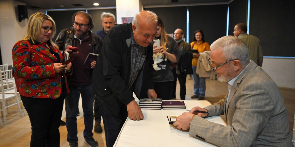 Kapalı bir mekanda düzenlenen imza gününde, gri ekose ceketli ve gözlüklü bir yazar masada oturmuş kitap imzalamaktadır. Yazarın önünde üst üste dizilmiş kitaplar bulunur. Yazarın karşısında duran, üzerinde kareli gömlek ve siyah ceket olan yaşlı bir adam, yazarın imzaladığı kitabı dikkatle izlemektedir. Etrafta ellerinde telefonları ve kitapları olan, etkinliği takip eden bir grup katılımcı bulunmaktadır.