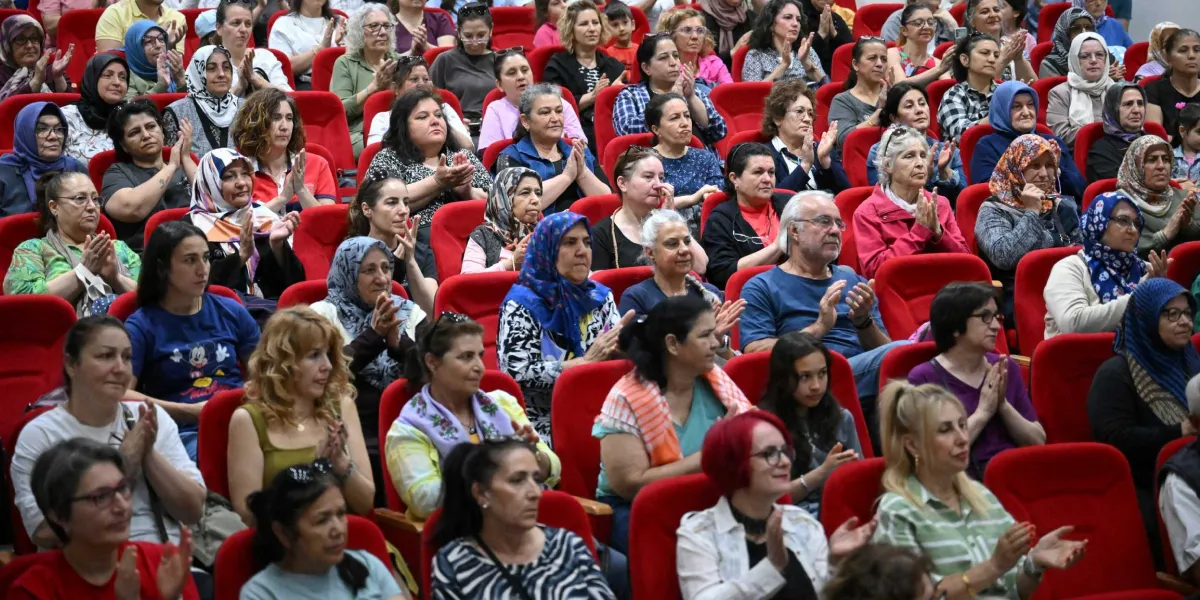 Konferans salonunda kırmızı koltuklarda oturan ve alkışlayan kalabalık bir kadın grubu. Farklı yaş ve giyim tarzlarına sahip kadınların bir kısmı başörtülü, bir kısmı ise günlük kıyafetler içinde dikkatle sahneye bakarak alkış tutuyor. Salonun genelinde odaklanmış ve katılımcı bir atmosfer hakim.