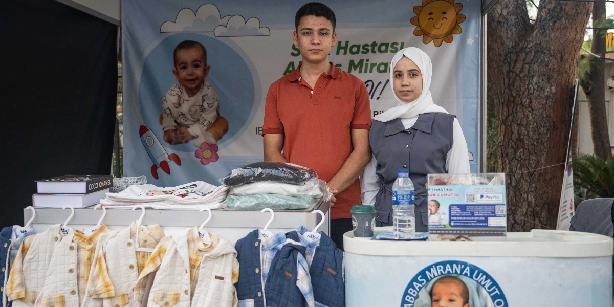 Bi Hayat Fest kapsamında, SMA hastası Abbas Miran için düzenlenen yardım standında duran genç bir erkek ve başörtülü genç bir kadın. Arka planda Abbas Miran'ın fotoğrafının yer aldığı bir afiş bulunurken, ön taraftaki stantta satılmak üzere dizilmiş çocuk yelekleri, kıyafetler ve üzerinde bağış bilgileri ile QR kod bulunan bir bilgilendirme kartı yer alıyor.