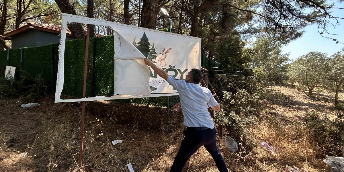 Üniformalı bir zabıta memuru, ağaçlık bir alanda bulunan metal çerçeveli bir reklam tabelasının üzerindeki brandayı söküyor. Tabelanın üzerinde tavşan figürü ve 