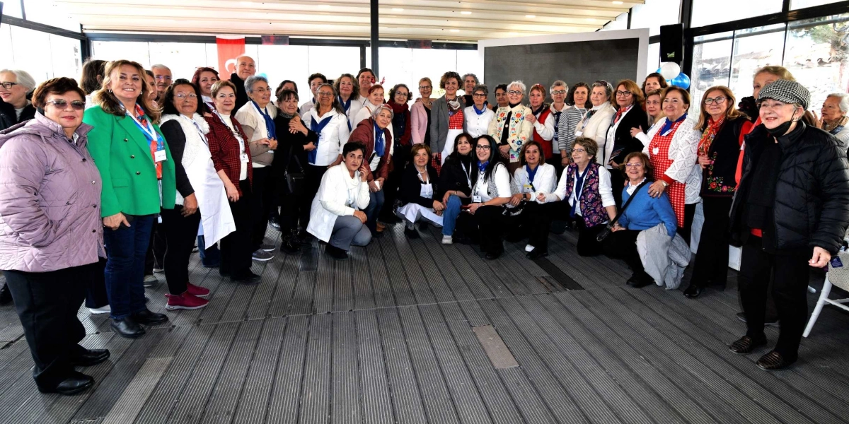 Kapalı bir mekanda, ahşap zemin üzerinde bir araya gelmiş, çoğunluğu kadınlardan oluşan kalabalık bir grup, gülümseyerek kameraya poz veriyor. Bazı katılımcılar ayakta dururken, bir kısmı ön sırada oturuyor. Katılımcıların birçoğu boyunlarına mavi fular takmış durumda ve genel olarak günlük, şık kıyafetler giyiyorlar. Arka planda cam duvarlar ve bir etkinlik alanı görülüyor.