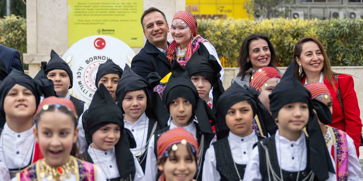 Bornova Belediye Başkanı Mustafa İduğ, 23 Nisan Ulusal Egemenlik ve Çocuk Bayramı etkinlikleri kapsamında geleneksel kıyafetler giymiş bir grup çocukla birlikte poz veriyor. Başkan İduğ, kucağında yöresel kıyafetli bir kız çocuğuyla gülümseyerek dururken, çevresinde siyah başlıklar ve beyaz gömlekler giymiş diğer çocuklar yer alıyor. Arka planda Bornova Belediyesi logosunun bulunduğu bir anıt ve iki kadın yetkili bulunuyor.