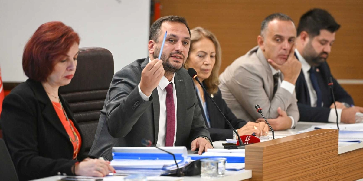 Bornova Belediyesi meclis toplantısında, kürsüde konuşma yapan bir yetkili elinde kalemle bir noktaya işaret ederken görülüyor. Toplantı masasında kendisiyle birlikte üç kişi daha oturuyor ve önlerinde dosyalar ile mikrofonlar bulunuyor.