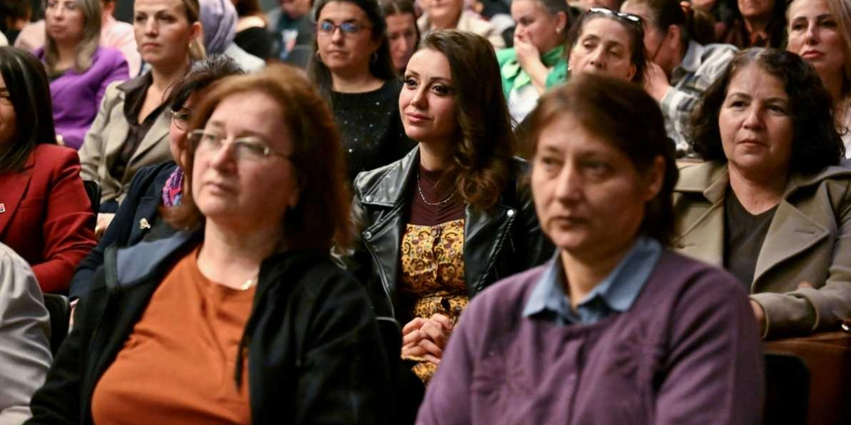 Kapalı bir salonda, bir sunumu veya konuşmayı dikkatle dinleyen, çoğunluğu kadınlardan oluşan kalabalık bir izleyici grubu. Ön planda, kiremit rengi bluz ve siyah hırka giyen gözlüklü bir kadın ile yanında mor hırkalı bir başka kadın yer alıyor. Arka planda ise farklı kıyafetler içinde, odaklanmış bir şekilde ileriye bakan çeşitli yaş gruplarından kadınlar bulunuyor.