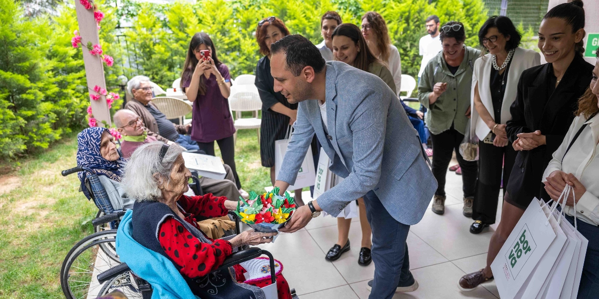 Açık havada, yeşillikler içindeki bir mekanda, açık mavi ceketli bir erkek, tekerlekli sandalyede oturan yaşlı bir kadına Anneler Günü anısına renkli kağıtlardan yapılmış çiçek buketi hediye ediyor. Çevredeki diğer katılımcılar gülümseyerek bu anı izliyor ve ellerinde üzerinde Bornova Belediyesi logosu bulunan hediye çantaları taşıyorlar.